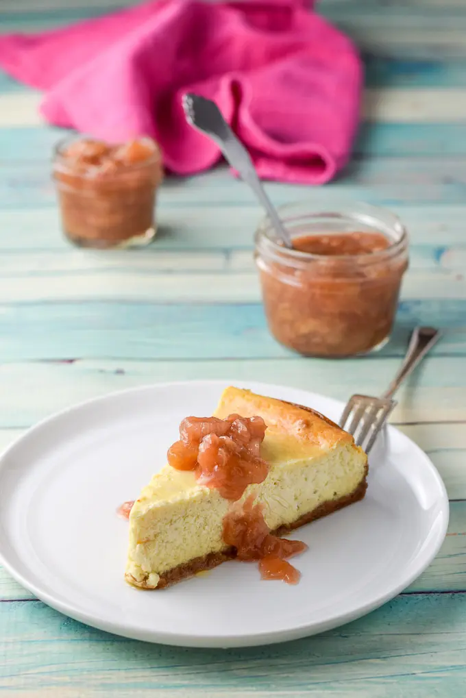 Slice of cheesecake dribbled with rhubarb with two jars full of sauce in back