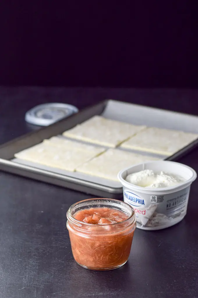 Rhubarb sauce and container of cream cheese in front of a jelly roll pan with four puff pastry squares on a sheet of parchment paper