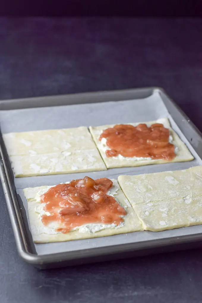 A jelly roll pan with four puff pastry squares on a sheet of parchment paper, with a layer of rhubarb sauce on top of a layer of cream cheese