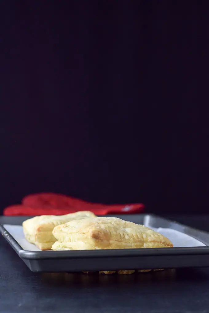 Vertical view of two puff pastry squares stuffed with cream cheese and rhubarb sauce baked to a golden brown on a jelly roll pan