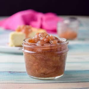 A small jar filled with the sauce with cheesecake in the background along with another jar and napkin