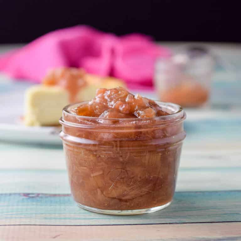 A small jar filled with the sauce with cheesecake in the background along with another jar and napkin