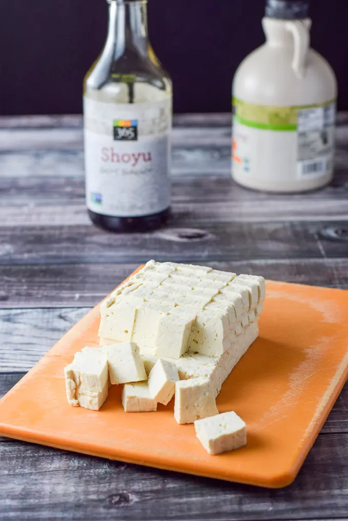 Tofu cut in cubes on a orange cutting board with soy sauce and maple syrup in the background