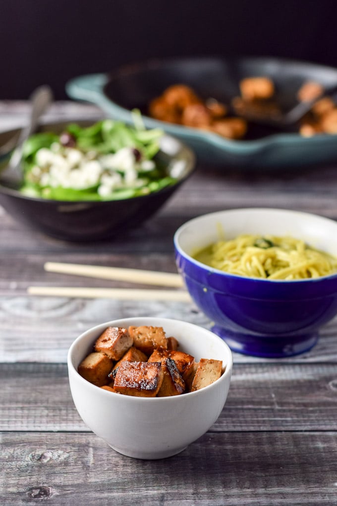 A small bowl filled with browned tofu with a blue bowl with noodles, a salad and the rest of the tofu in a pan in the background
