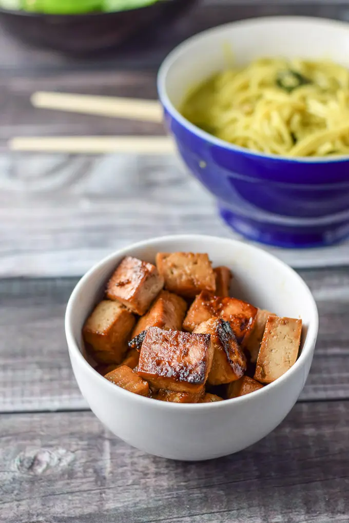 A white bowl with sautéed tofu with a bowl of noodles in the background