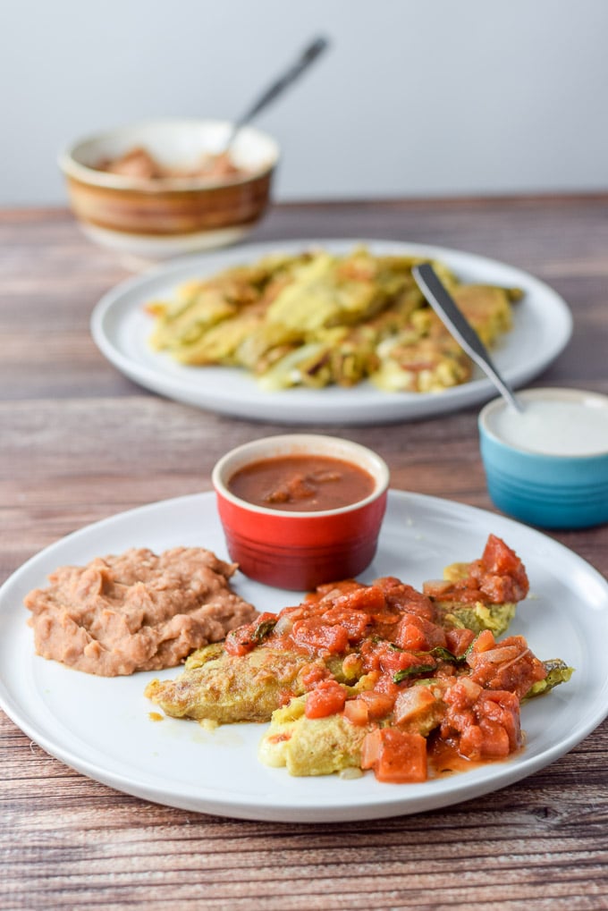 A white plate with two chile rellenos, beans and enchilada sauce. There is a plate filled with rellenos in the background