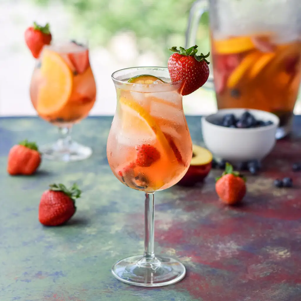 Two tulip glasses filled with fruit, ice and sangria with a pitcher of it in the background along with fruit - square