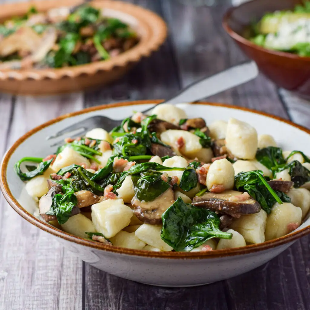 A white bowl with the gnocchi dish in it with a plate with the vegetable in - square