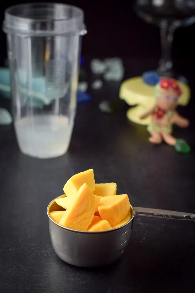 Mangos in a measuring cup in front of the blender container filled with the alcohol
