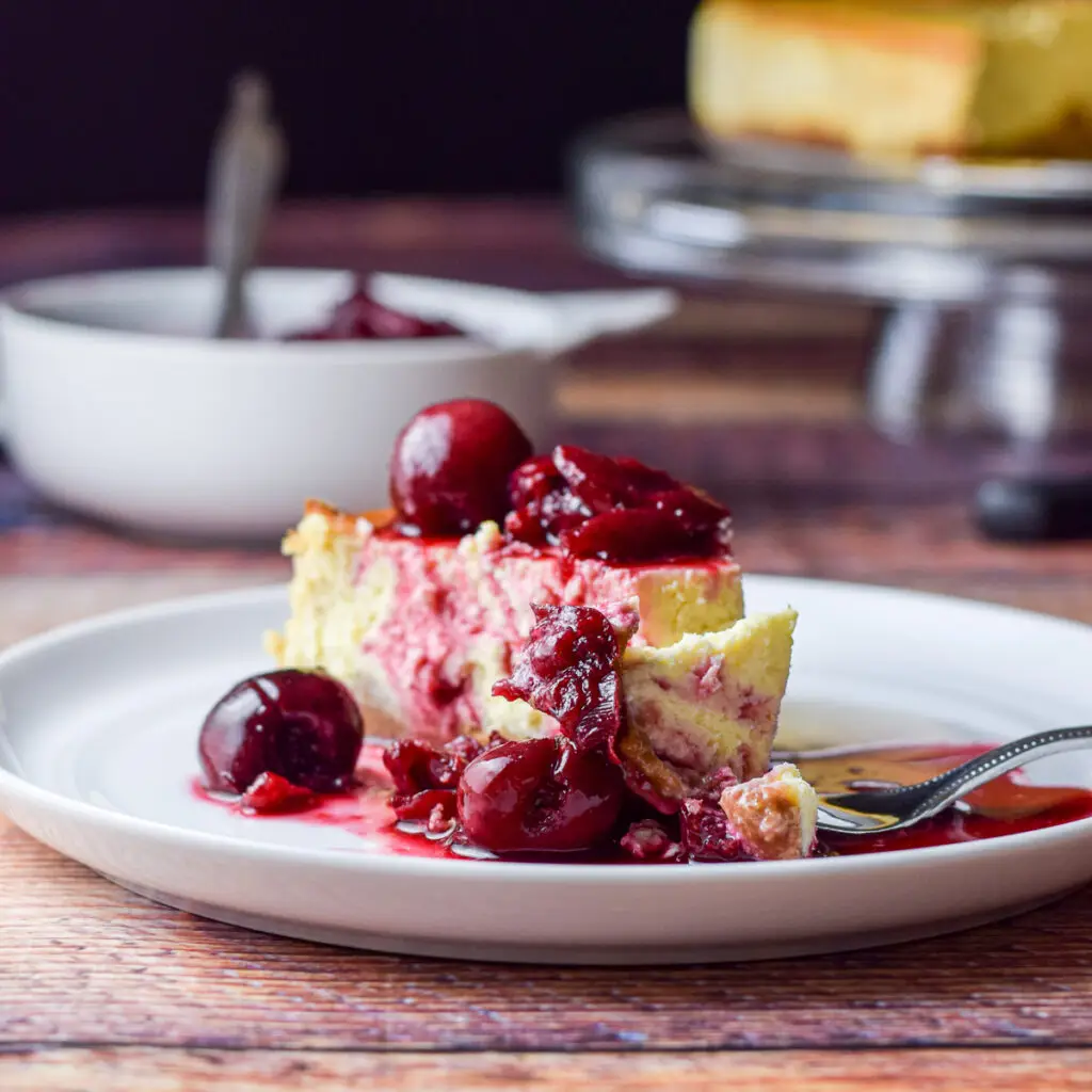 A small plate with the cheesecake covered with cherries and a fork with a bite laying on the plate - square