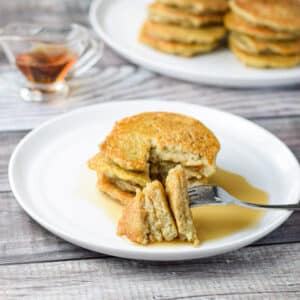 A fork with cut pancakes on it with a plate filled with them and maple syrup in the background - square