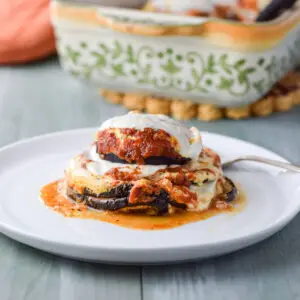 A close up of a vertical photo of a white plate with eggplant stacks on it with the baking dish in the background - square