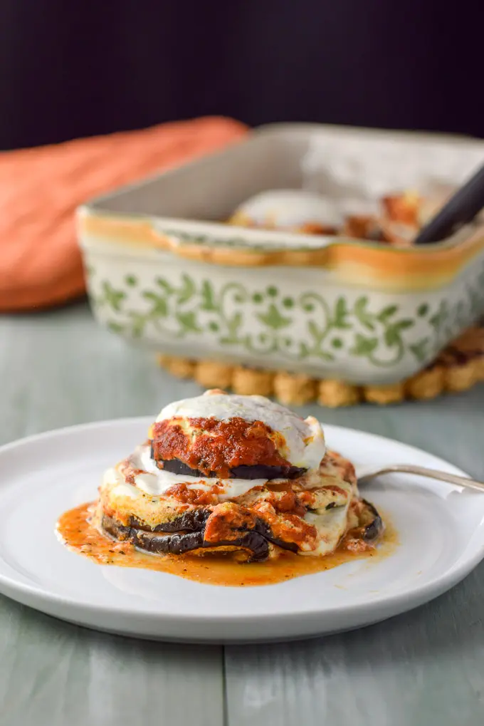 A close up of a vertical photo of a white plate with eggplant stacks on it with the baking dish in the background