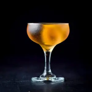 A coupe glass filled with amber, reddish liquid and a lemon twist - square