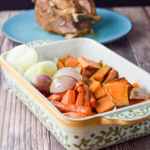 A baking dish filled with baked carrots, yams, shallots and onions. There is also a pork butt in the background