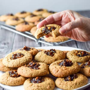A female hand holding a cookie with a bite taken out it. There is a plate of cookies and a wire rack in the background - square