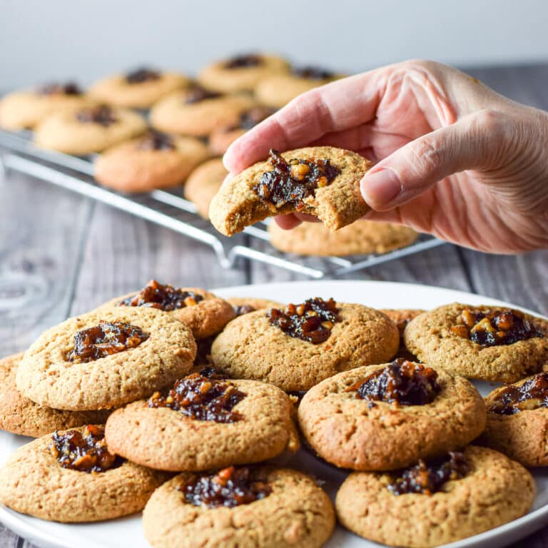 A female hand holding a cookie with a bite taken out it. There is a plate of cookies and a wire rack in the background - square