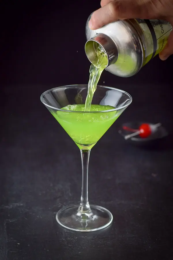 A hand pouring the green cocktail into a martini glass with a maraschino cherry is in the background