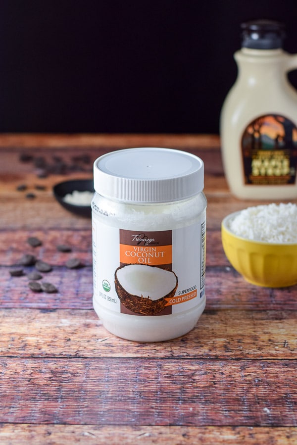 Container of coconut oil, shredded coconut and flour and a bottle of maple syrup in the background