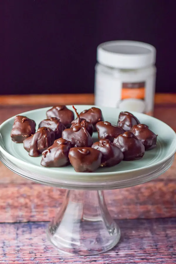 A plateful of chocolate coconut candies sitting on a cake stand with a container of coconut oil in the background.