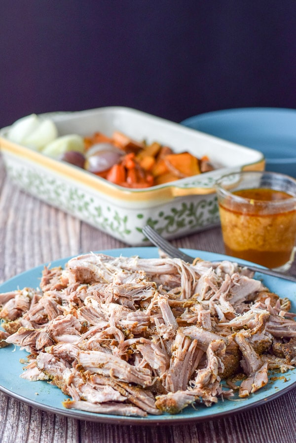A big blue platter with the pork, shredded and some pork sauce in a pitcher and veggies in a baking dish in the background