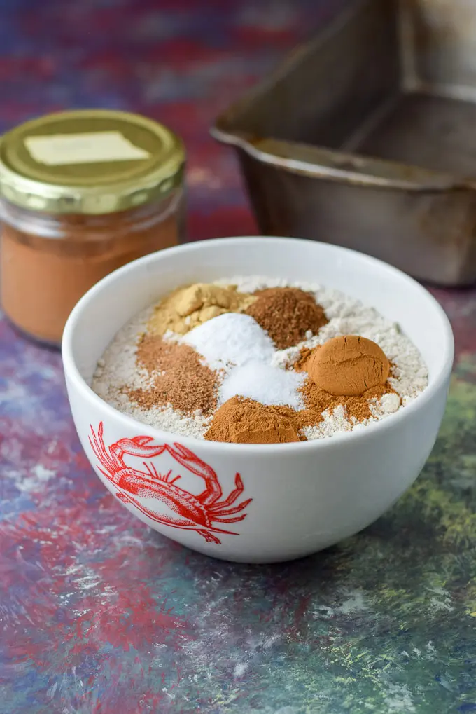 Cinnamon, ginger, cloves and nutmeg in a bowl of flour with a big jar of cinnamon in the background and a bread pan