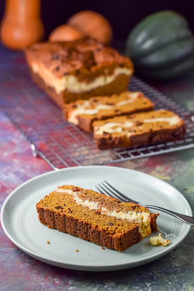 A plated piece of pumpkin bread with the cheesecake layer showing. There is the sliced bread in the background