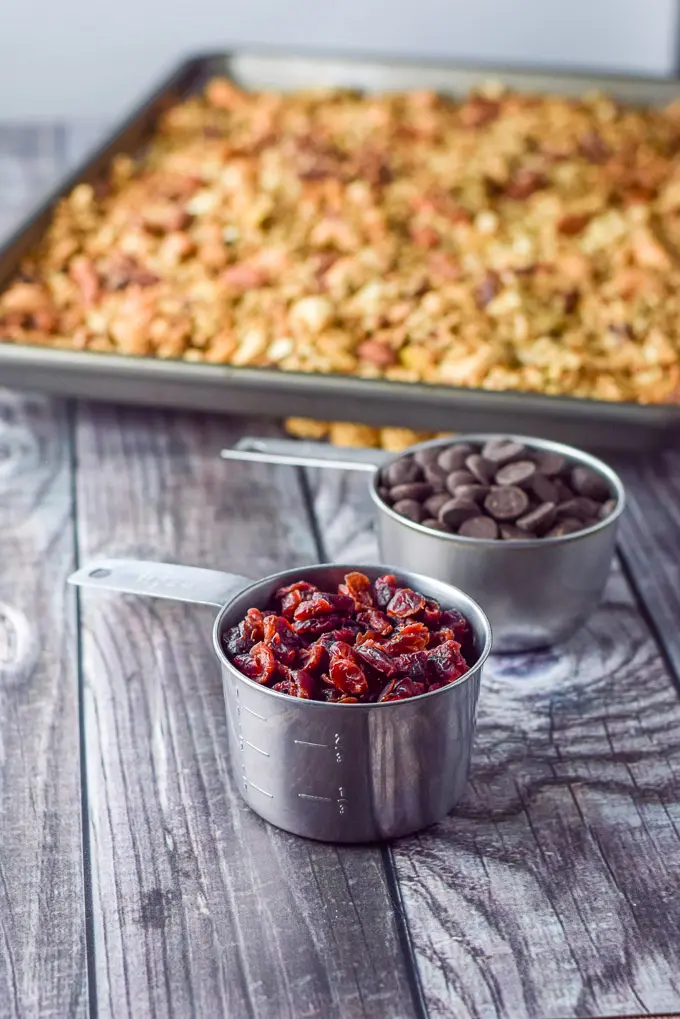 Two measuring cups with cranberries and chocolate in them and the pan of granola in the background