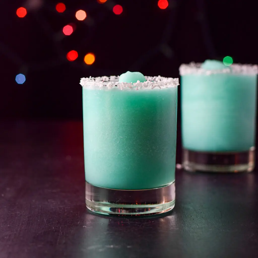 Vertical view of two glasses with coconut rims filled with a blue cocktail and lights in the background - square
