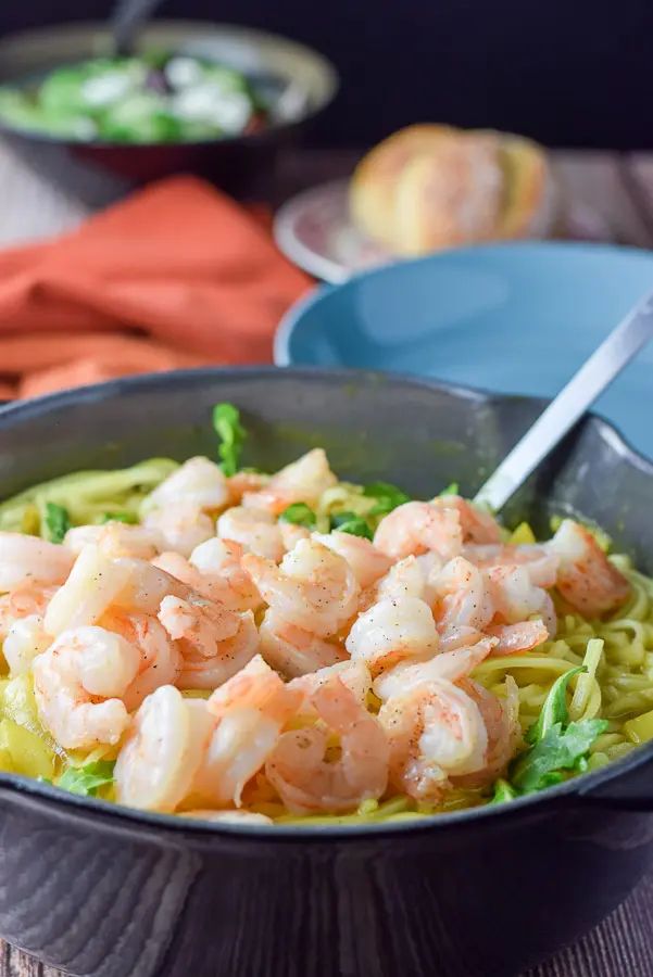 Shrimp piled on the noodles in a pan with a plate, roll and salad in the background