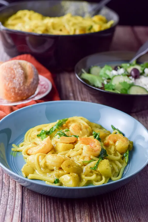 A blue bowl with shrimp on noodles in front of the pan, salad and roll