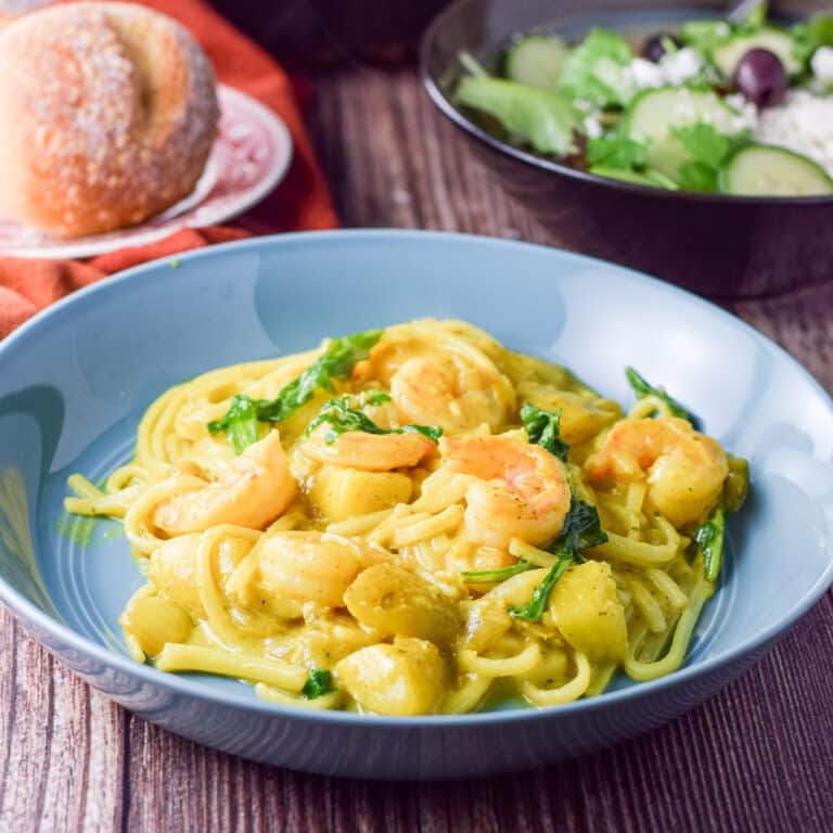 A blue plate with noodles and shrimp with a salad and roll in the background - square