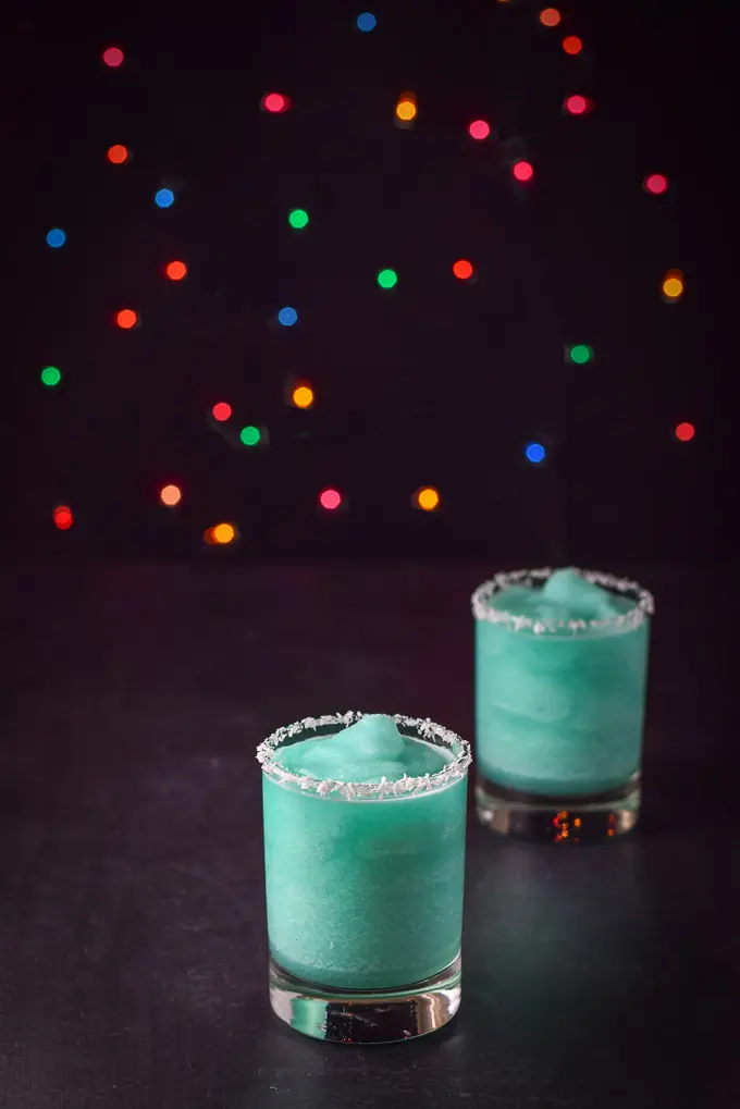 Two Jack Frost cocktails with coconut rims on a black table and colorful lights behind them