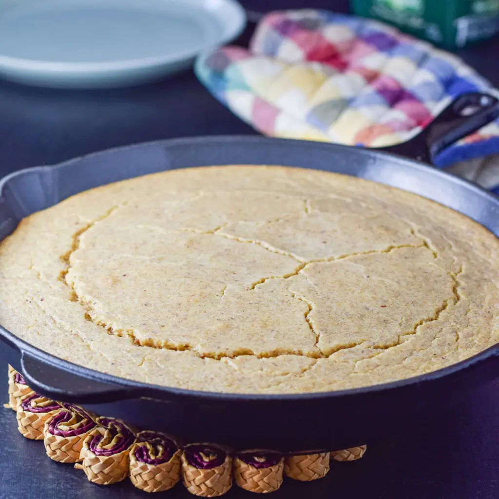 A cast iron pan filled with cornbread. There is a oven mitt and plate behind it - square