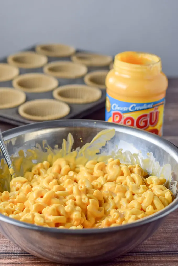 The elbow macaroni mixed with the cheese sauce in a metal bowl with muffin tins in the background