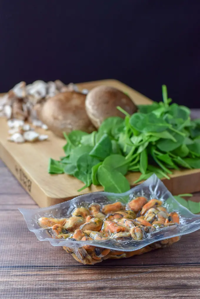 Mussels in a package on the table and spinach and mushrooms on a wooden board in the background