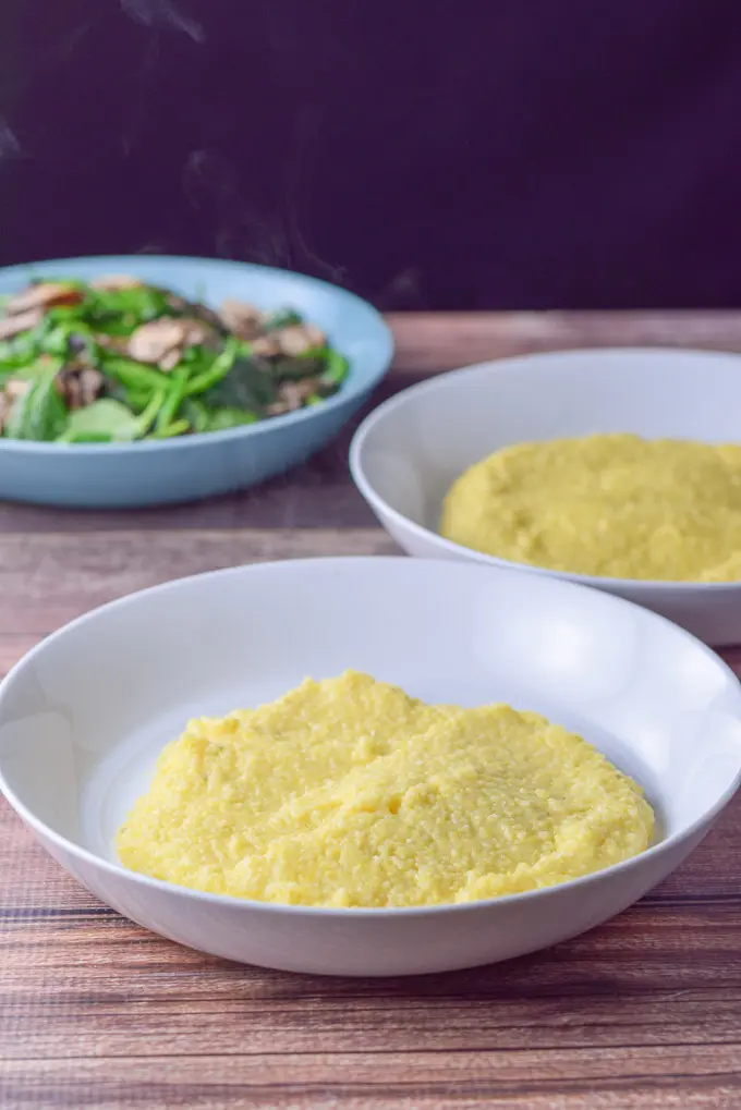 Polenta spooned in two bowls with some mushrooms and spinach in the background