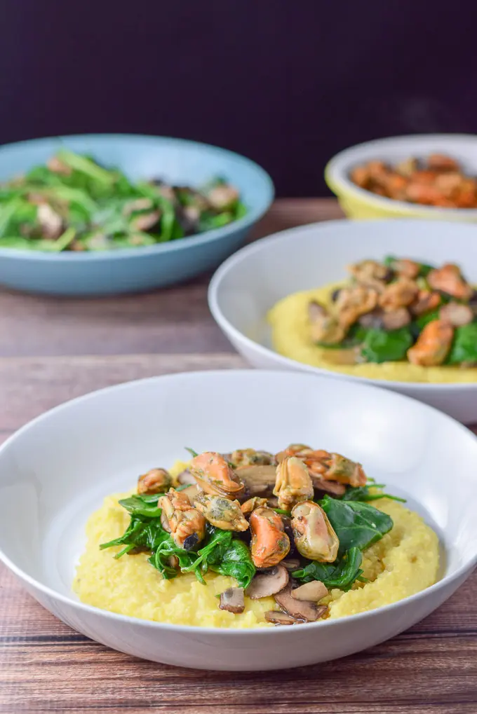 Two white shallow bowls of polenta topped with mushrooms, spinach and mussels