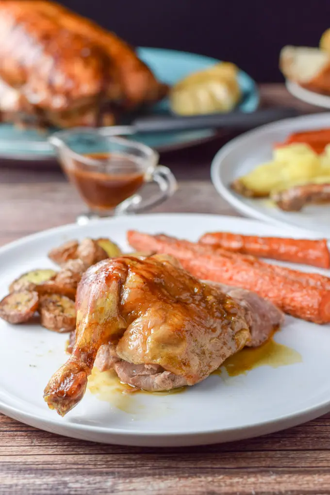 Close up of the duck leg quarter on the plate with carrots and potatoes. There is gravy and a duck on a platter in the background