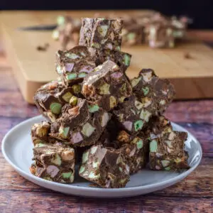 A square photo of fudge piled up on a plate with a more on a chopping block behind it