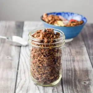 A pile of granola in a ball jar with a scoop and bowl in the background - square