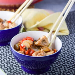Bowl of stir fry in a bowl with chop sticks in the bowl. There is a wok filled with the dish in the background - square