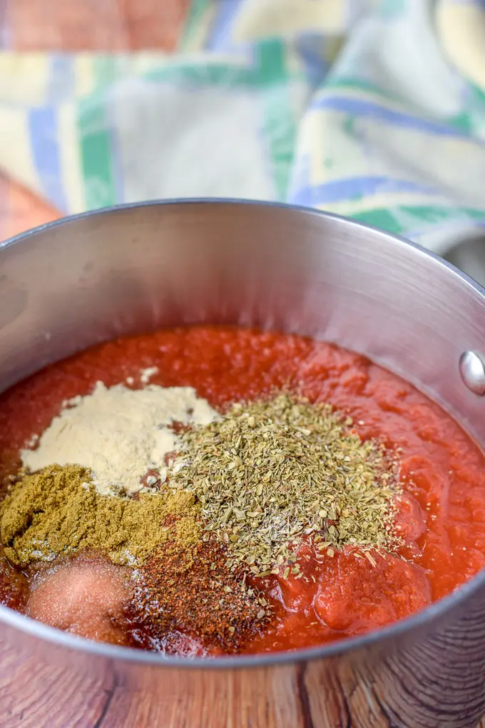 Crushed tomatoes in a pan with oregano, chili powder, cumin, salt and garlic powder added on top