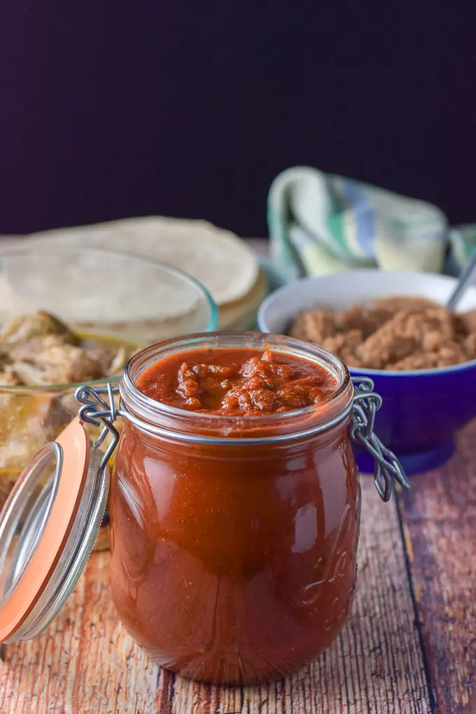 Red enchilada sauce in a jar with beans in the background along with pulled pork and whole wheat tortillas