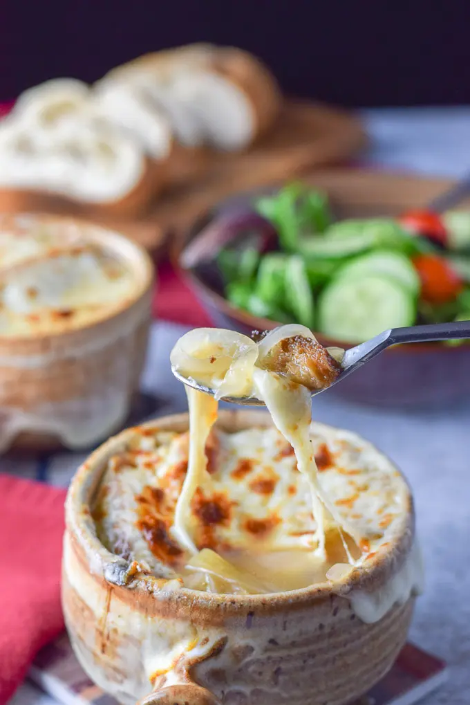 A cheesy spoonful of onion soup held over the crock with salad and bread in the background