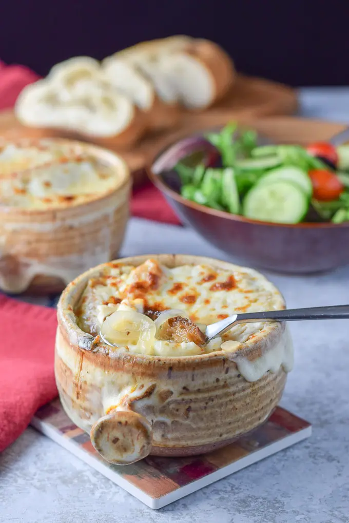 A spoon is in the crock of cheesy onion soup with a salad and sliced bread in the background