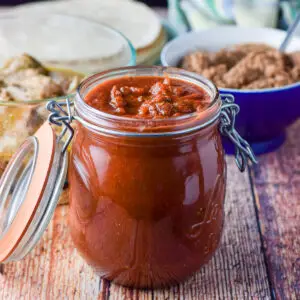 Red enchilada sauce in a jar with beans in the background along with pulled pork and tortillas - square