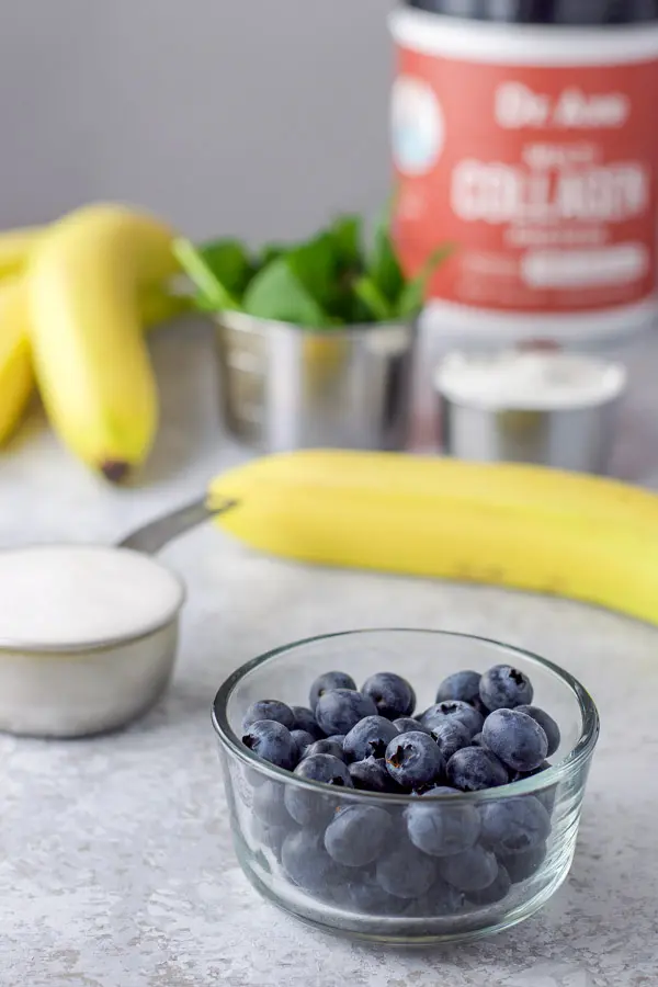 Blueberries in a glass bowl, milk, yogurt and spinach all in their own measuring cups and some bananas on a table