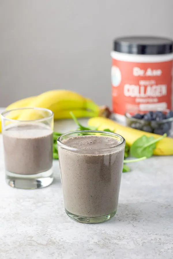 Two glasses filled with the spinach smoothie with collagen in the background as well as some blueberries and bananas