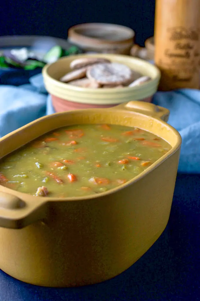A mustard colored casserole dish filled with pea soup. There are some pita bread in a bowl, a salad and a pepper mill in the background
